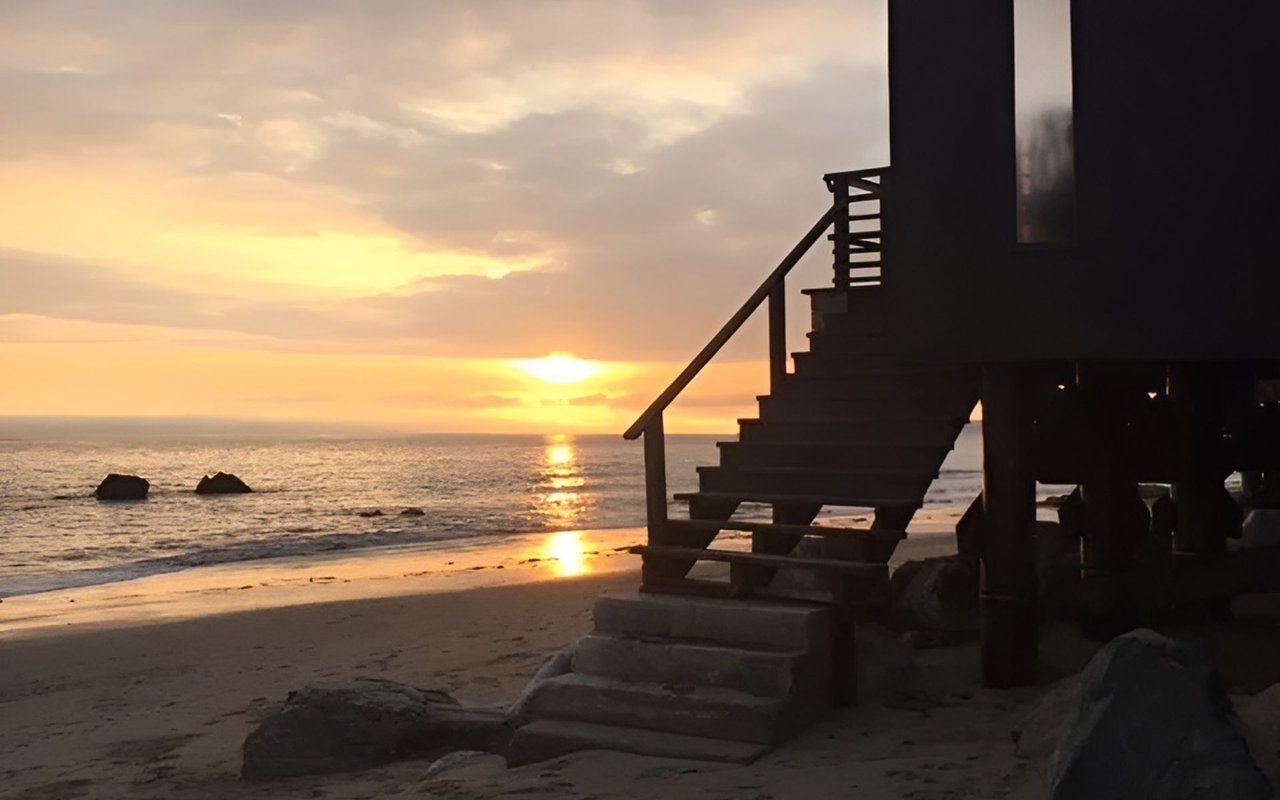 Beachfront house stairs overlook the ocean as the sun sets over the water.