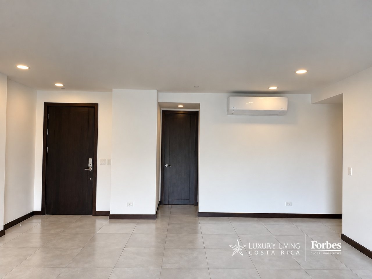 Vacant apartment living area with tiled floors, dark wood doors, and a wall-mounted air conditioner.