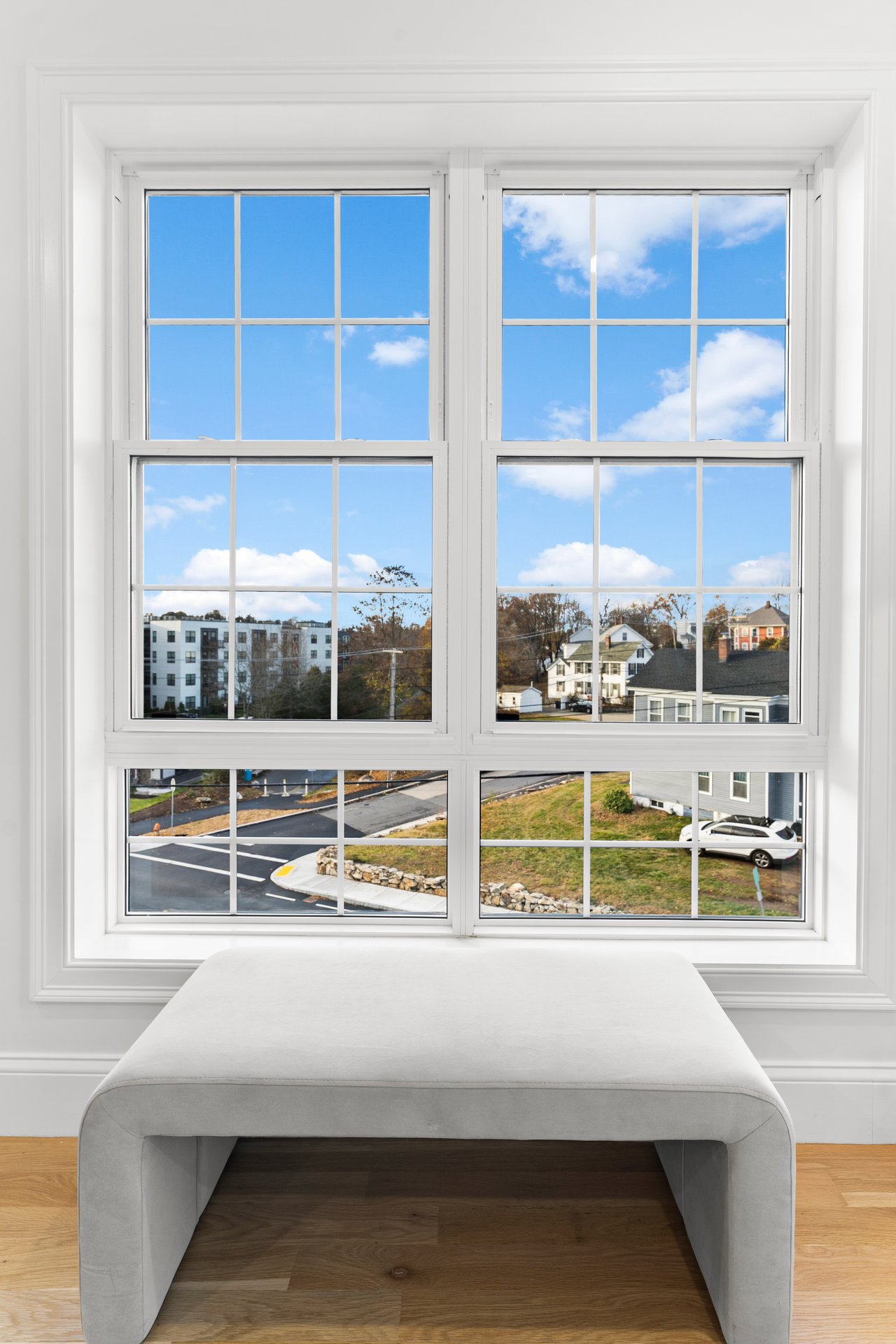 White window seat beneath a large divided-light window overlooking nearby homes and an autumn residential street.