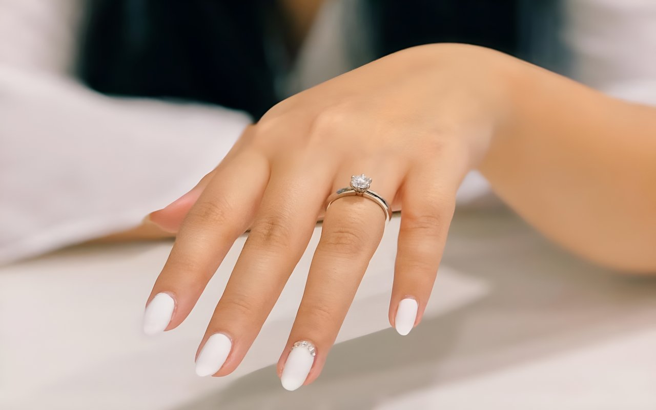 A hand with white nail polish and a diamond engagement ring on the ring finger.