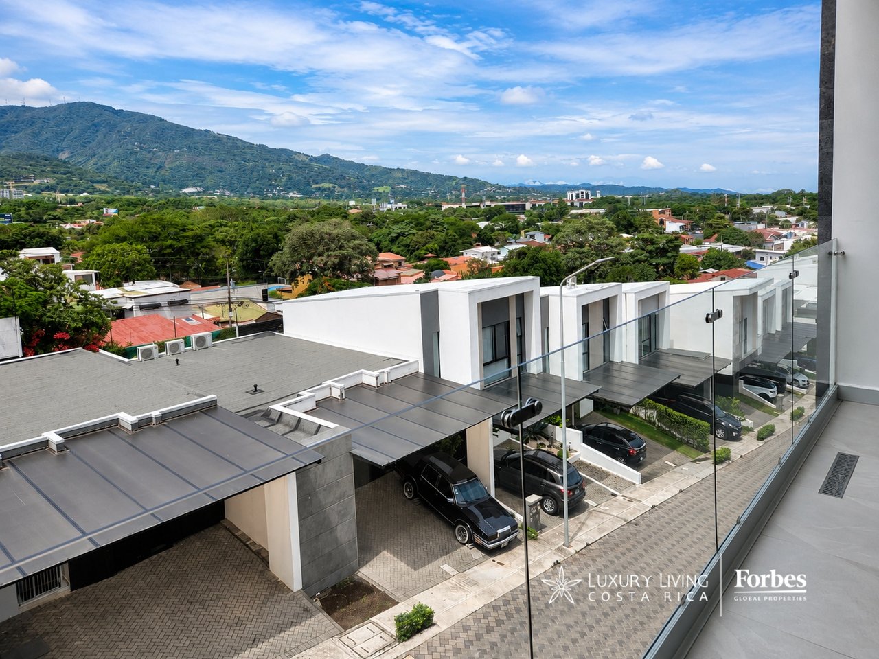 Modern residential buildings with glass balconies overlook a green neighborhood and mountain range.
