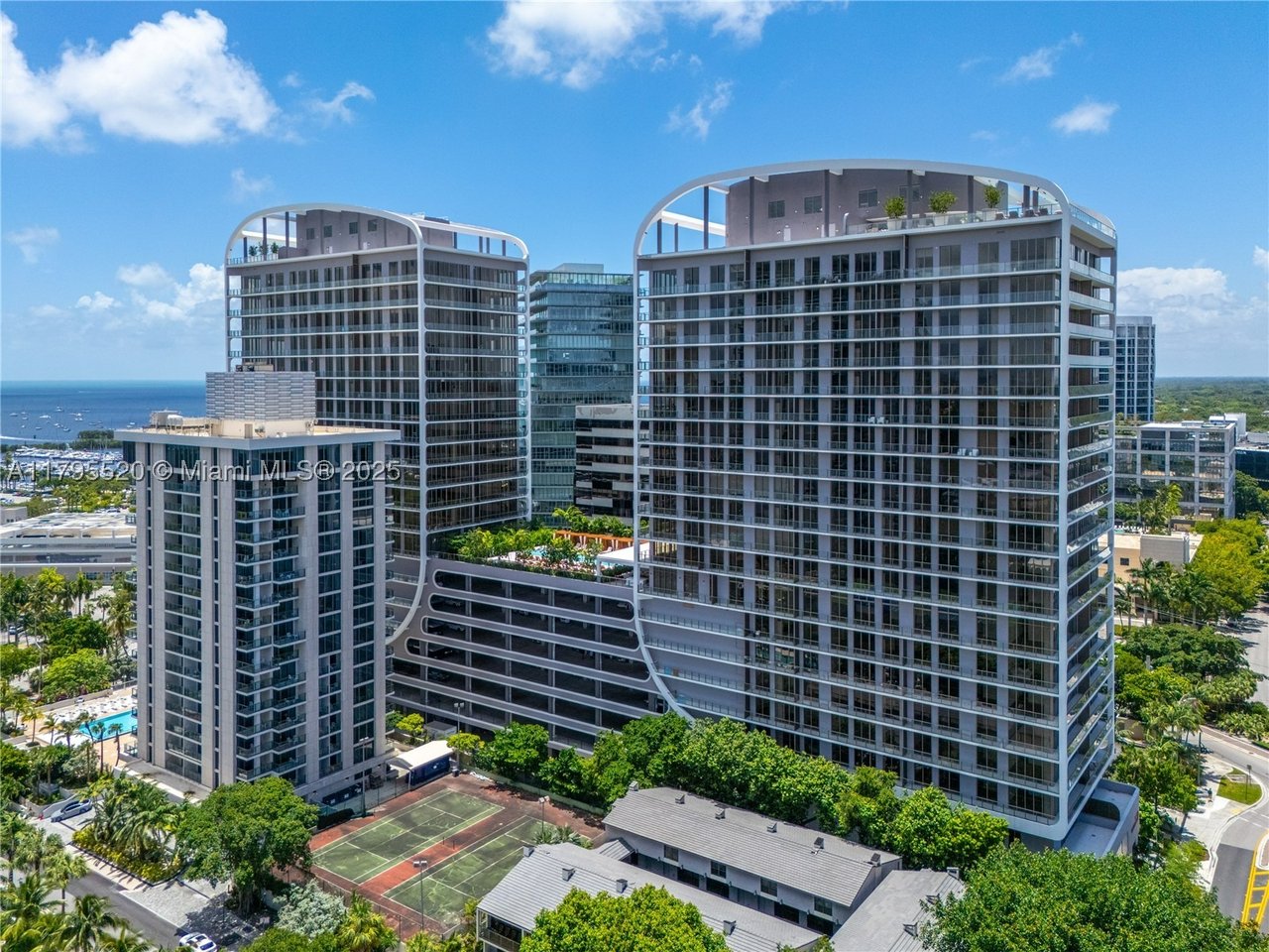 Two modern residential towers with curved rooftop frames, a tennis court, and ocean in the background.