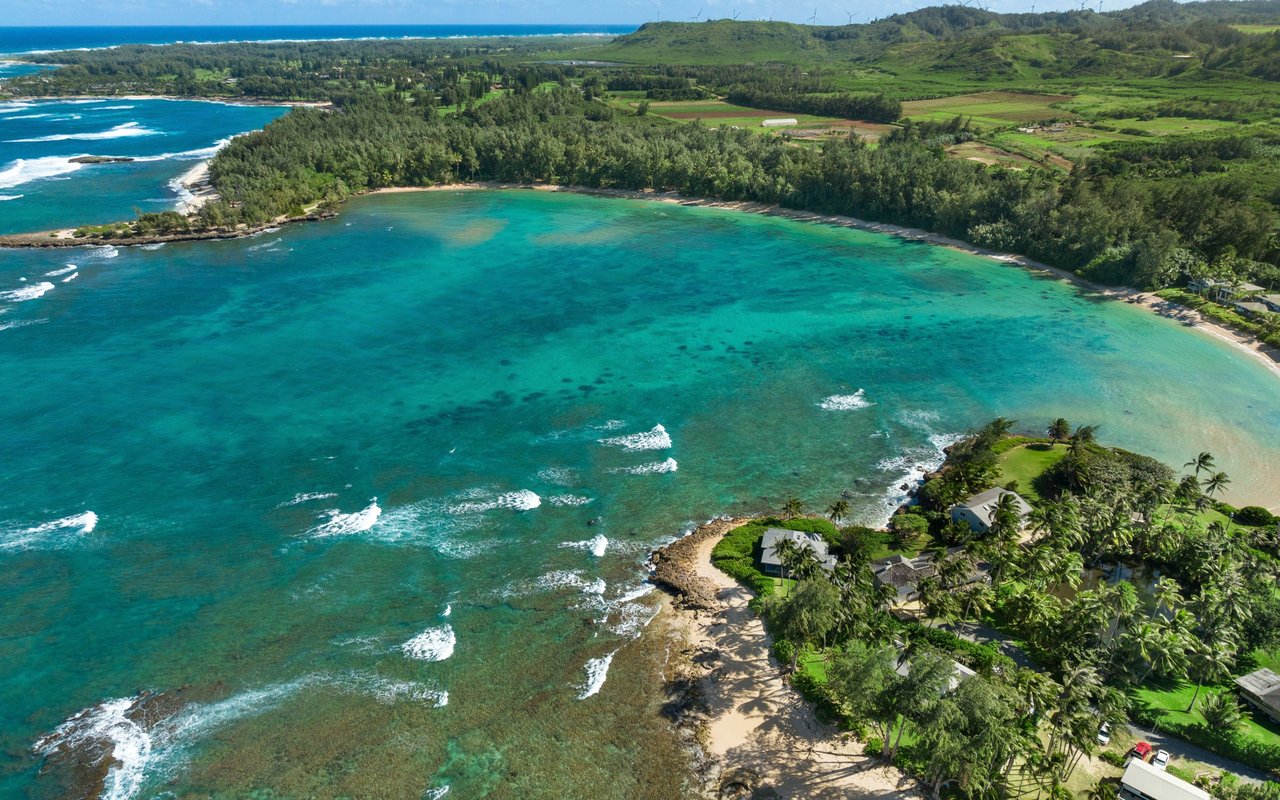 Aerial view of a turquoise Waikane coastline, with a sandy beach, wooded hills, and homes along the shore.