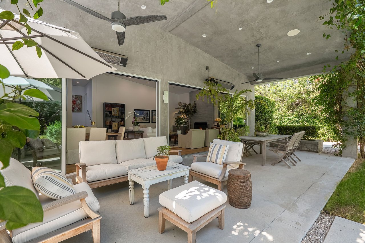 Covered patio with white-cushioned seating, a dining table, and ceiling fans beside open doors.