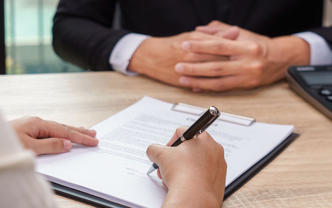 A person signs a document while another sits opposite with hands clasped.
