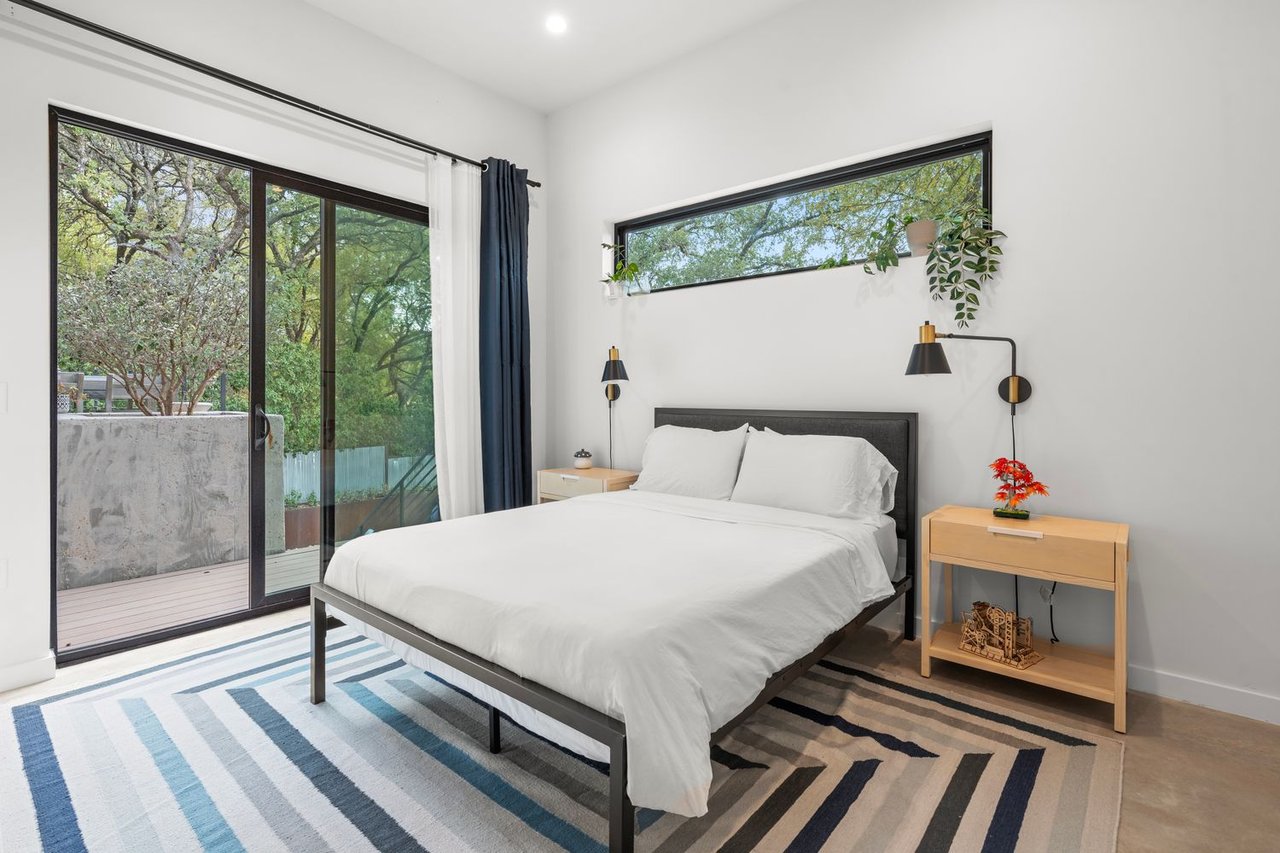 Staged bedroom with white bedding, striped rug, and sliding glass doors overlooking trees.
