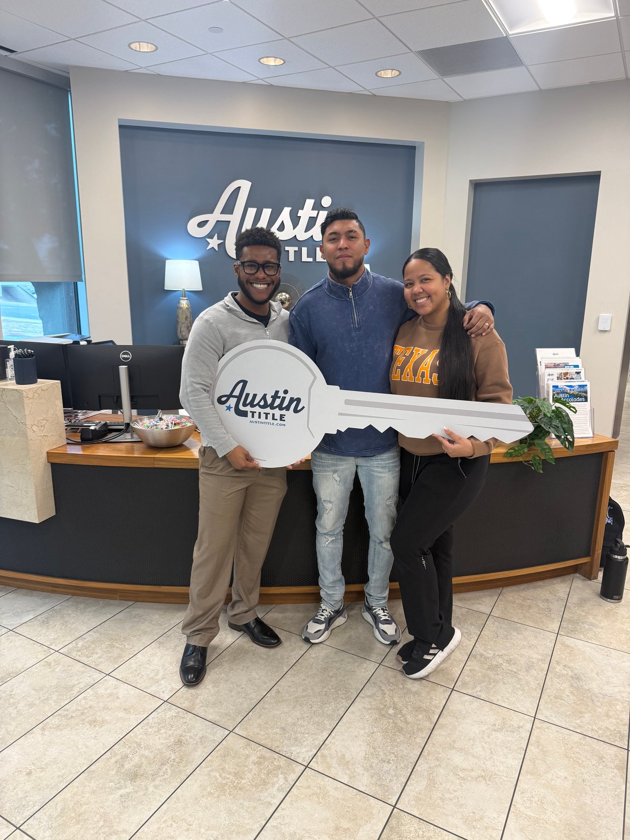 Three adults pose with an oversized Austin Title key sign at the office reception desk.