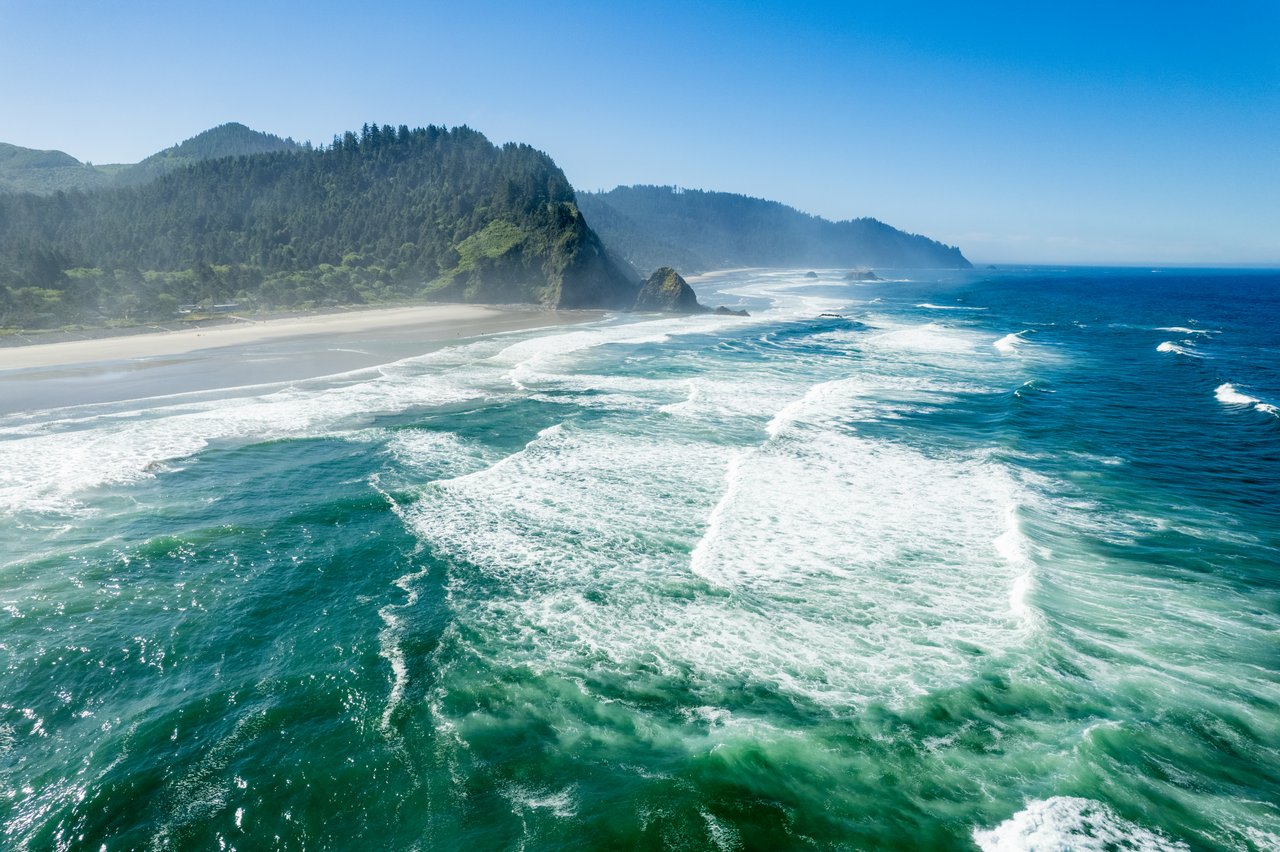 Pacific surf breaks along a sandy beach below forested coastal headlands.