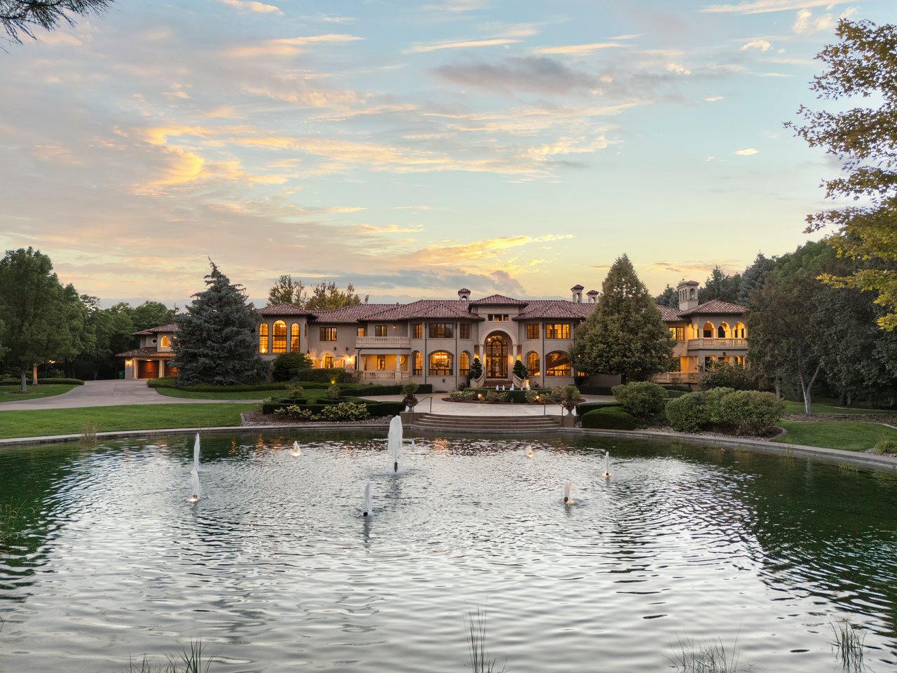 Large Mediterranean-style estate behind a fountain pond, with arched windows and landscaped grounds at sunset.