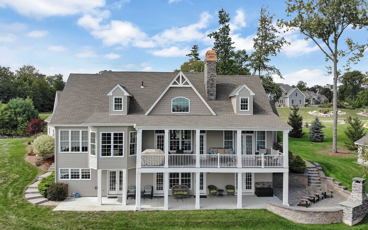 Gray two-story house with a wide covered porch, stone chimney, and furnished patio.