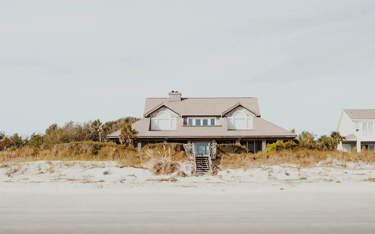 Raised beach house behind sandy dunes, with wooden stairs leading to its covered porch.