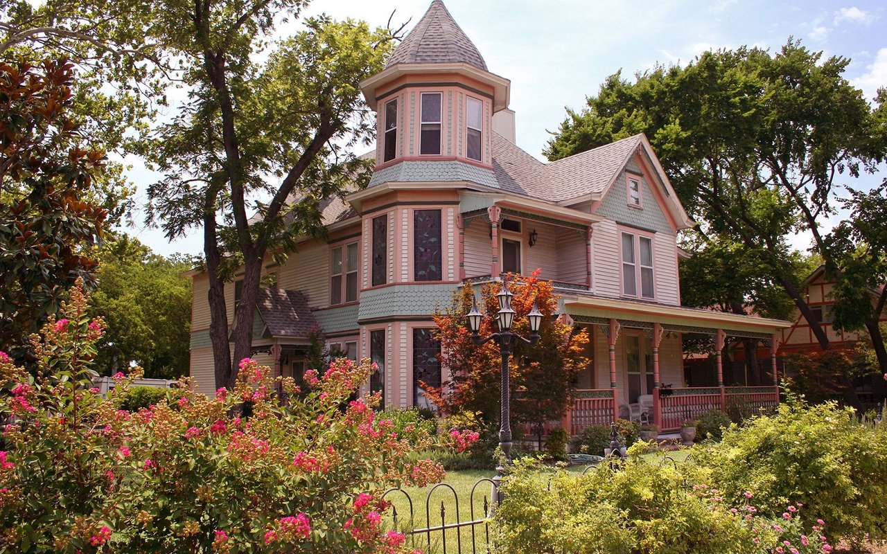 Pink Victorian-style house with a turret and wraparound porch amid flowering gardens.