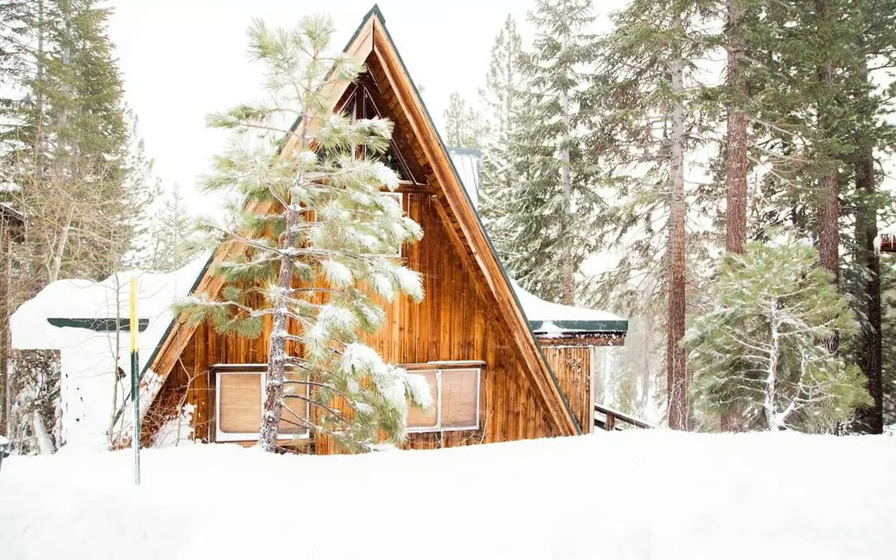 Wooden A-frame cabin surrounded by snow-covered evergreens.