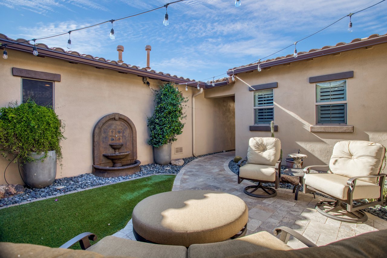 A stucco patio courtyard with string lights, a decorative wall fountain, potted greenery, and cushioned outdoor chairs around a paved seating area.
