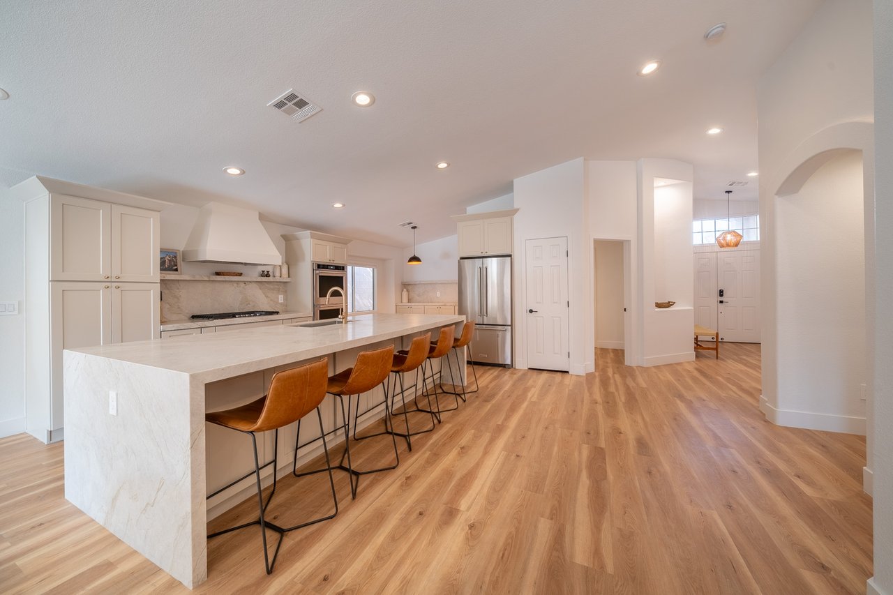 Open kitchen with a long stone island, leather stools, stainless appliances, and wood flooring.