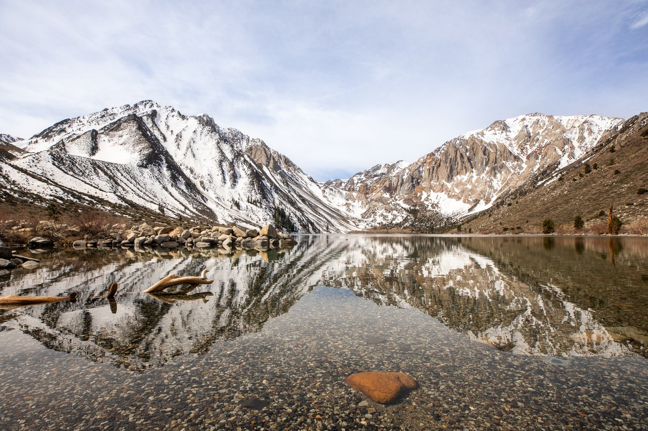 Alpine lake reflecting snow-dusted mountains, with a rocky shore and driftwood in the foreground.