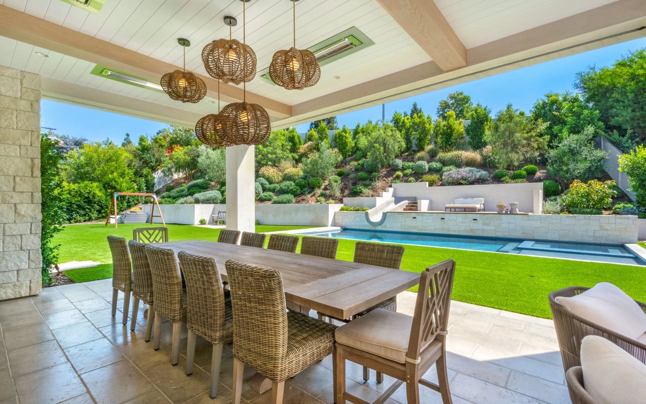 Covered patio with a long dining table overlooking a pool and landscaped hillside.