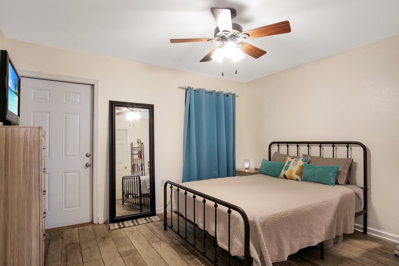 Cozy bedroom with a brown metal bed, beige quilt, and teal pillows. A full-length mirror and blue curtains add charm. Ceiling fan and wooden floor create warmth.