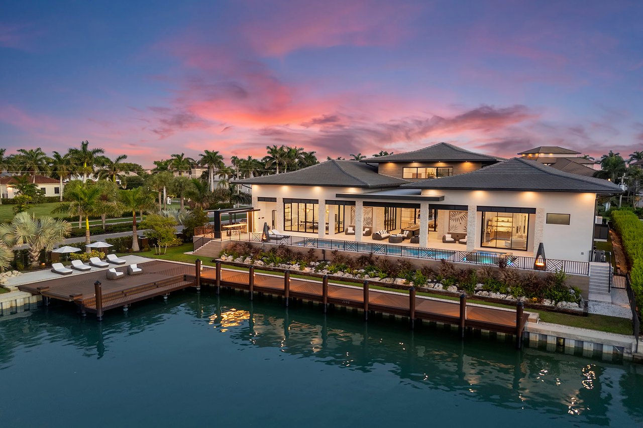 Modern waterfront home with a wooden dock and tropical landscaping beneath a pink dusk sky.