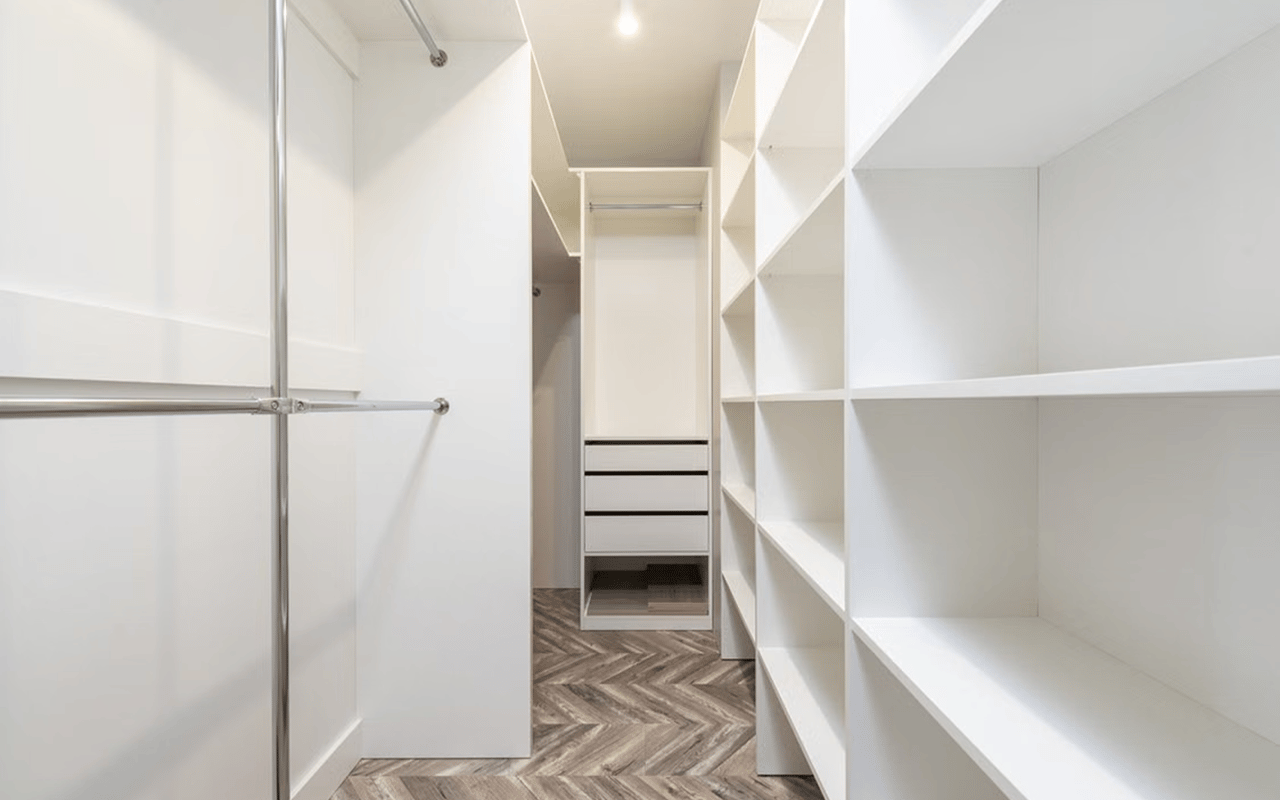 Walk-in closet with white built-in shelving, drawers, chrome hanging rods, and a herringbone wood floor.