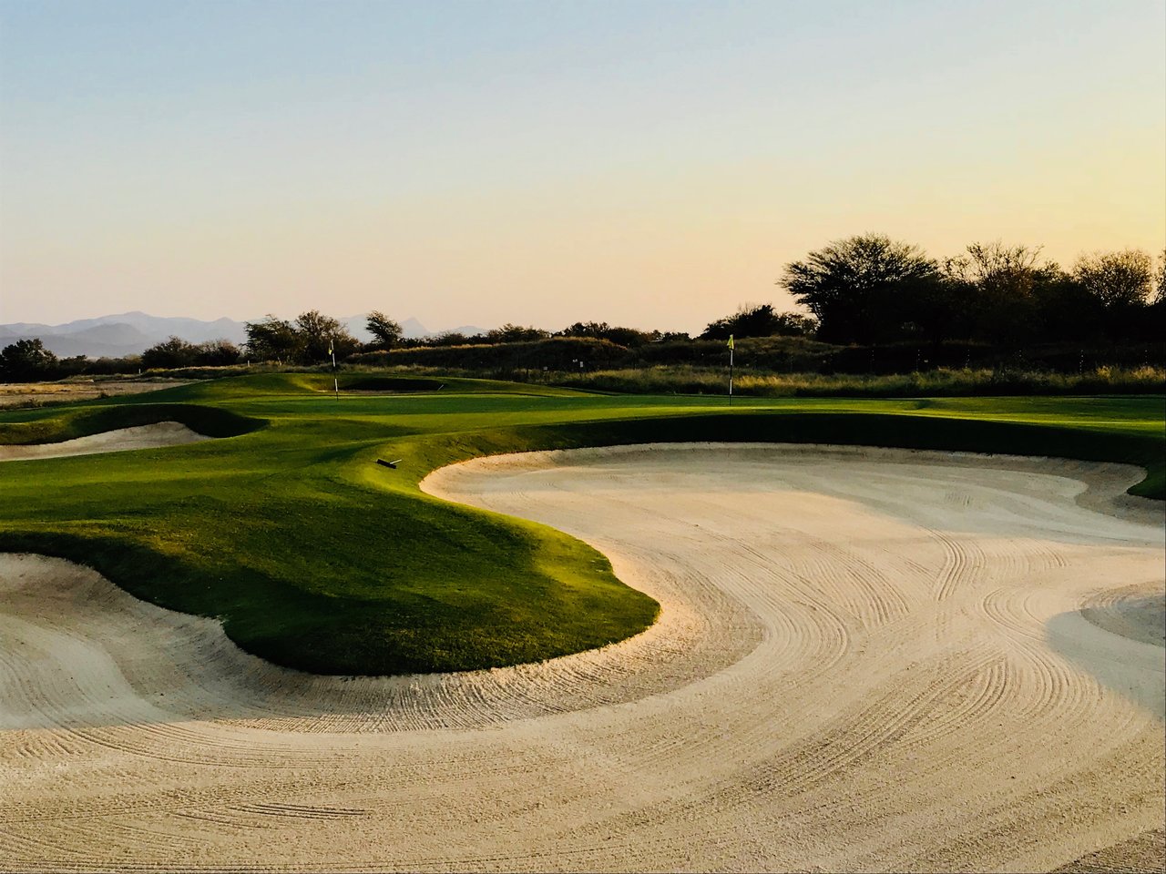Golf course green and flagsticks beside a large raked sand bunker at golden hour.