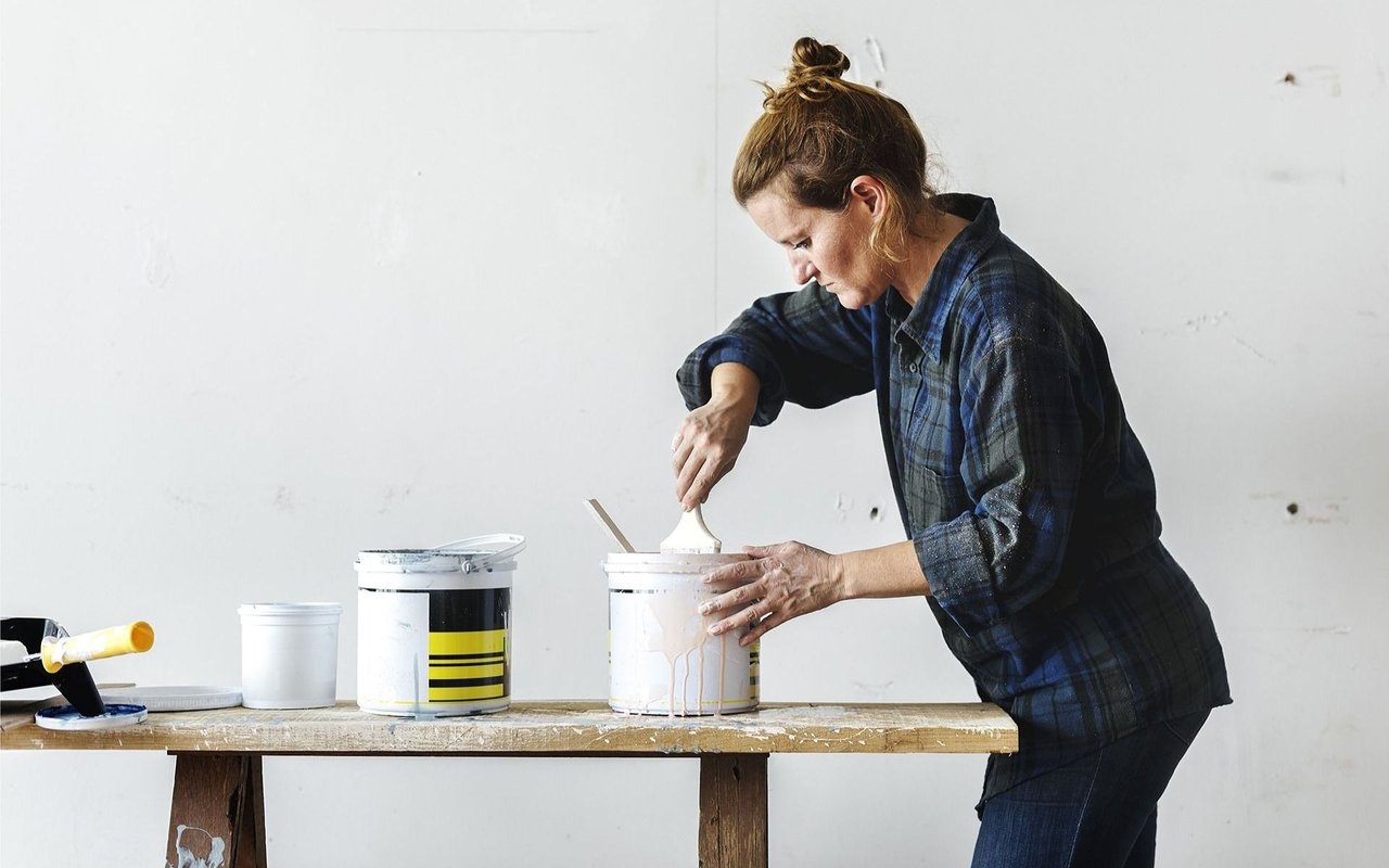 A woman stirs paint in a bucket at a workbench with paint supplies nearby.