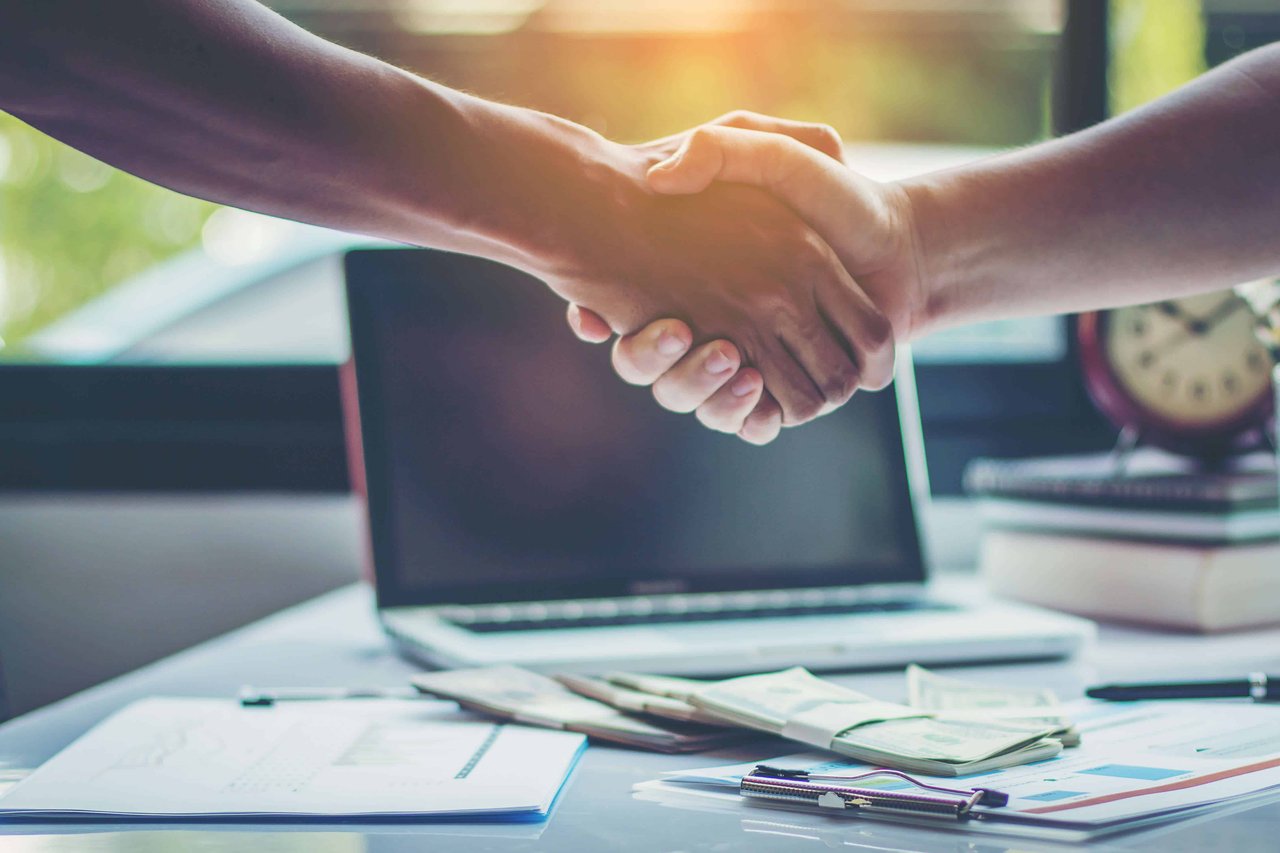 Two people shake hands across a desk scattered with paperwork beside an open laptop.
