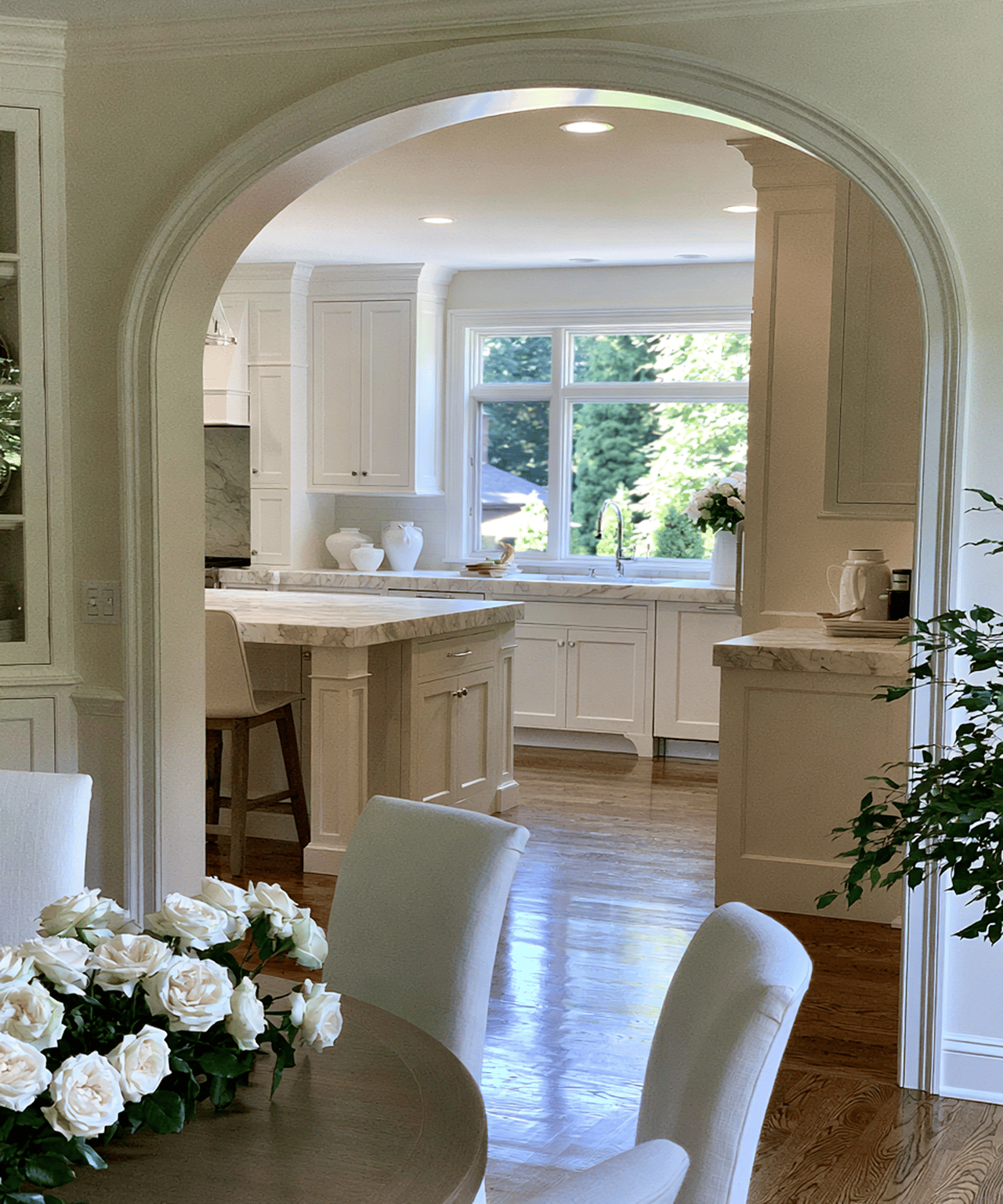 Bright white kitchen with marble counters and island, seen through an archway from a dining area with white roses.