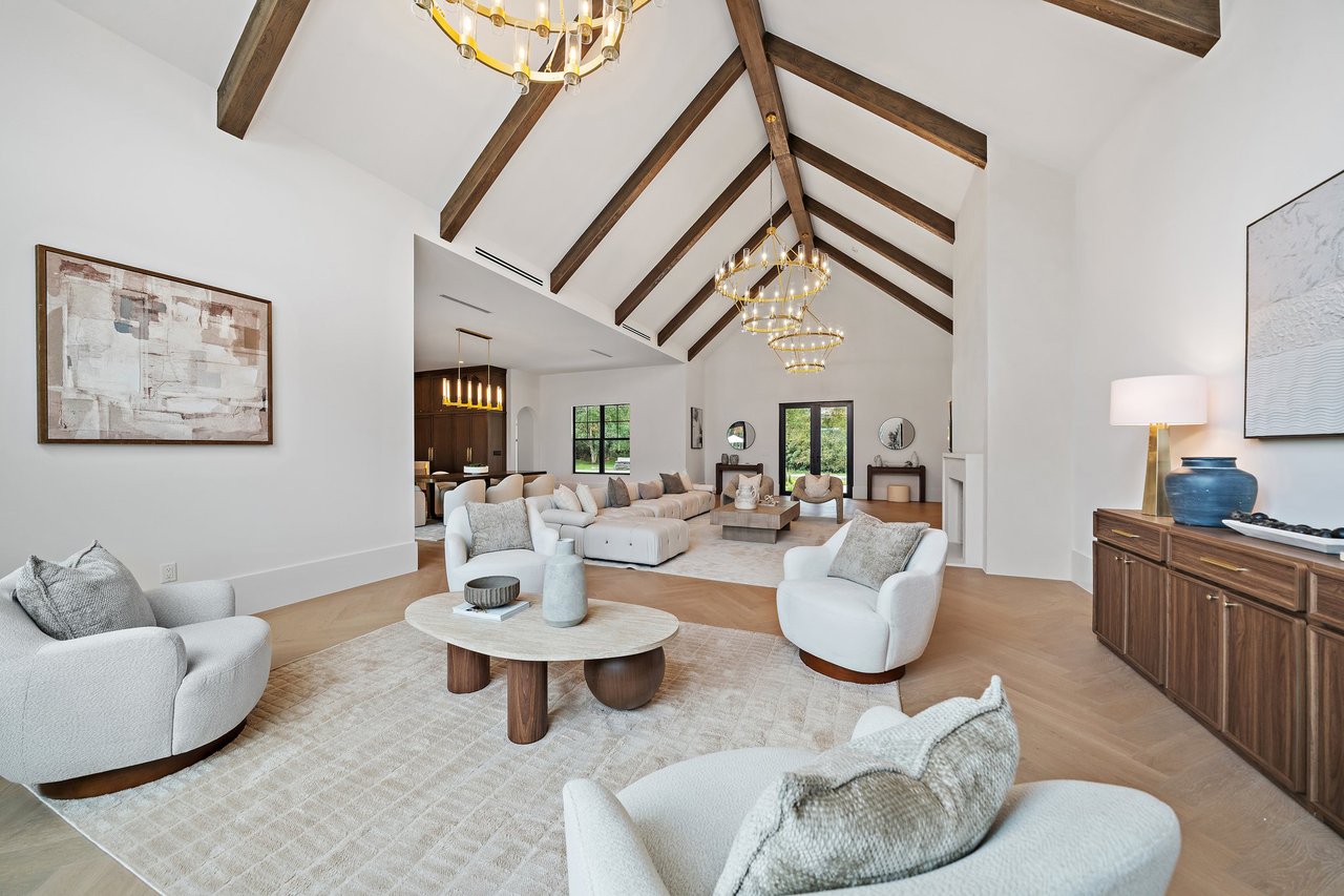 Vaulted great room with dark wood ceiling beams, gold chandeliers, and white seating.