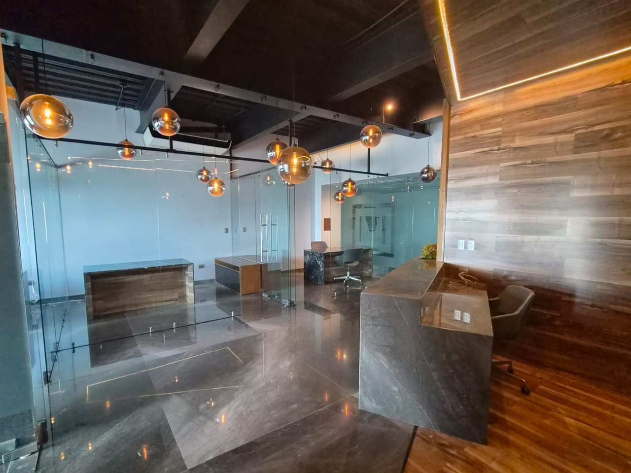Modern office reception with glass partitions, stone counters, wood paneling, and amber globe pendant lights.