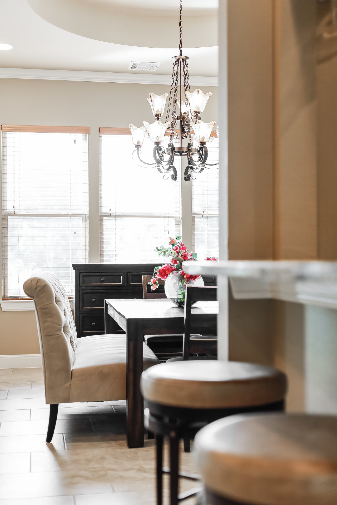 Dining room with a dark wood table, chandelier, bright windows, and a vase of red flowers.