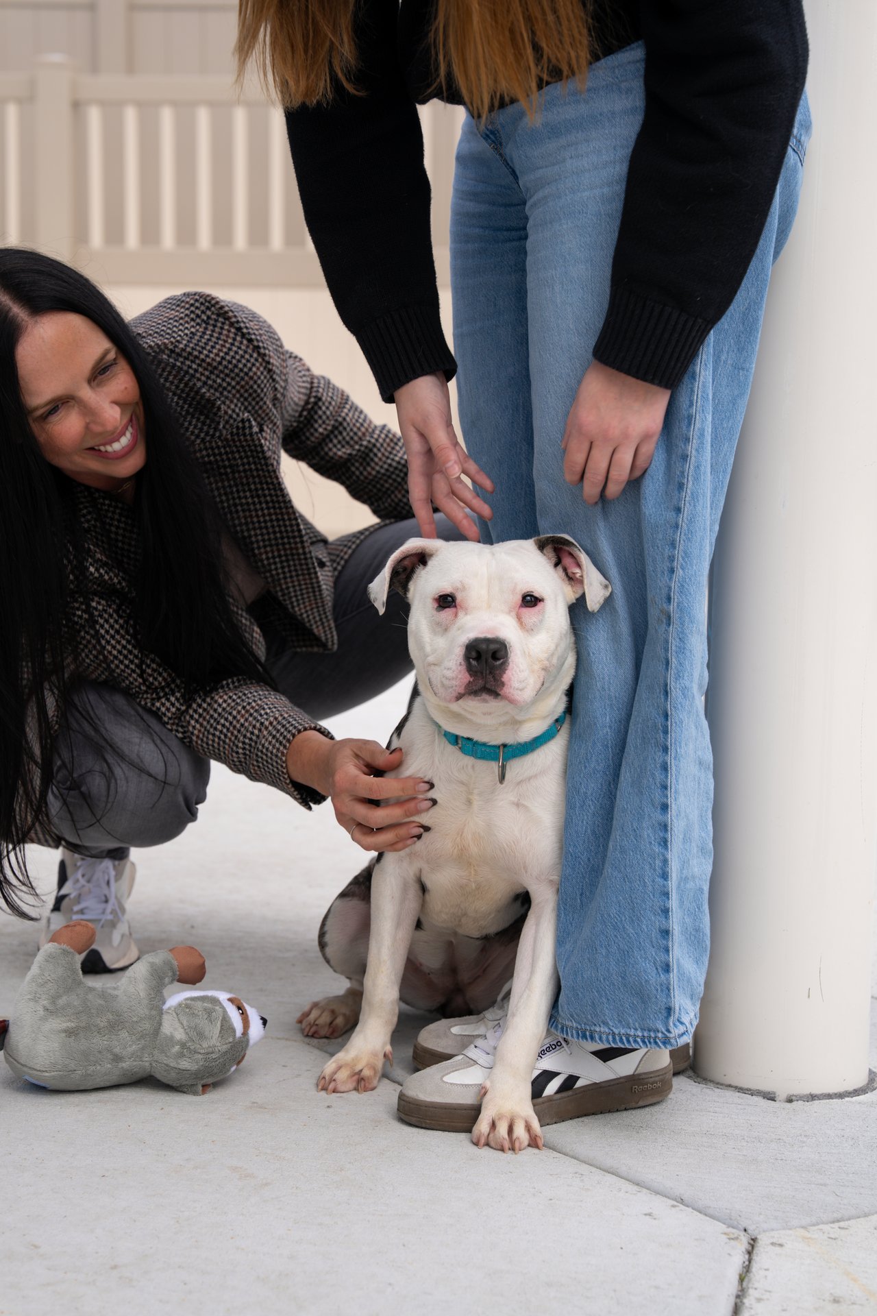 A woman and another visitor pet a seated white dog beside a plush toy.