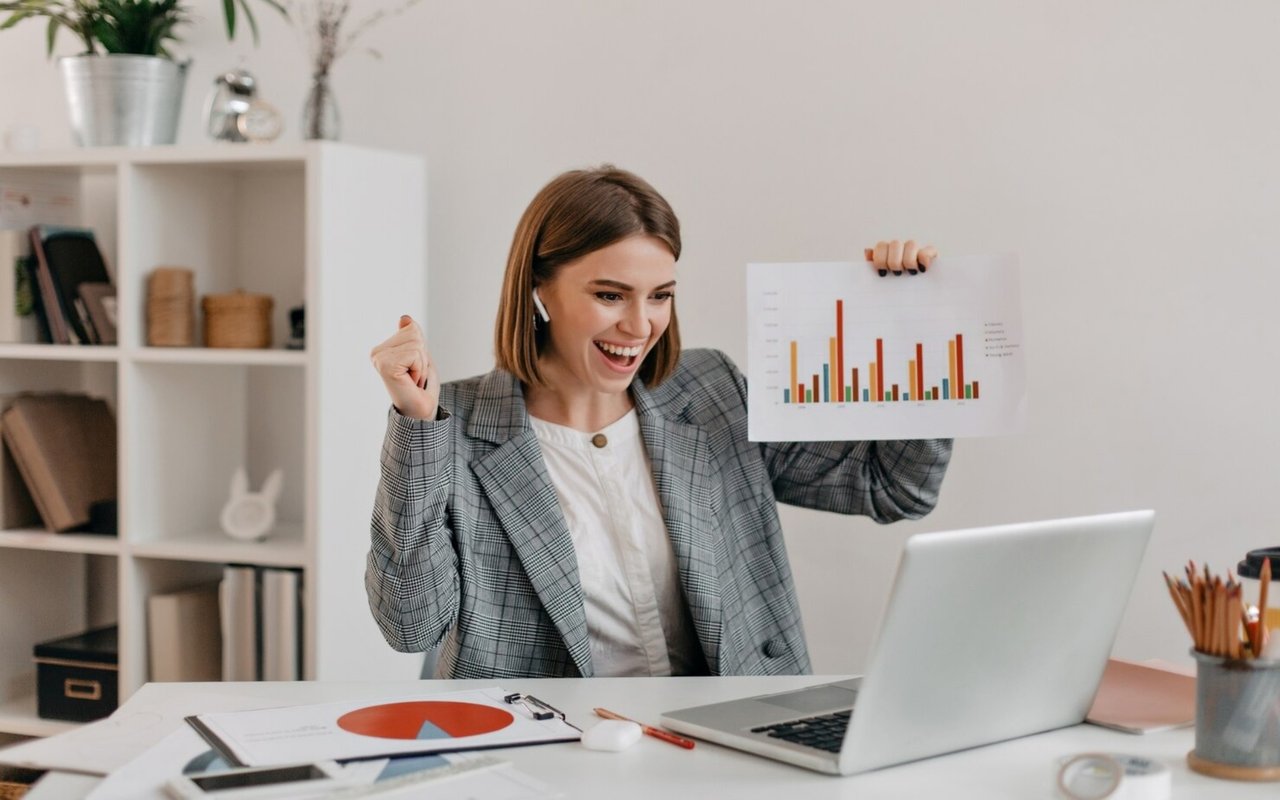 Woman celebrates while holding up a bar chart beside her laptop.
