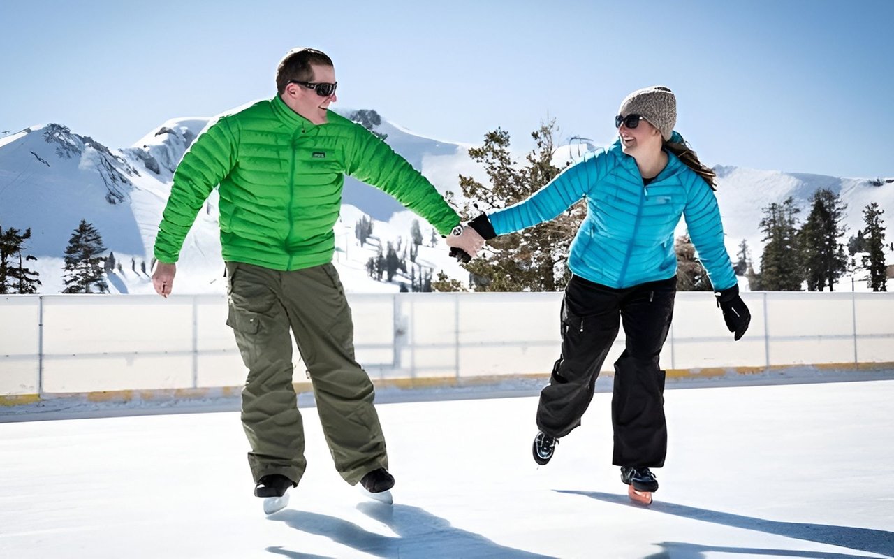 Two adults skate hand in hand on an outdoor rink, with snowy mountains behind them.