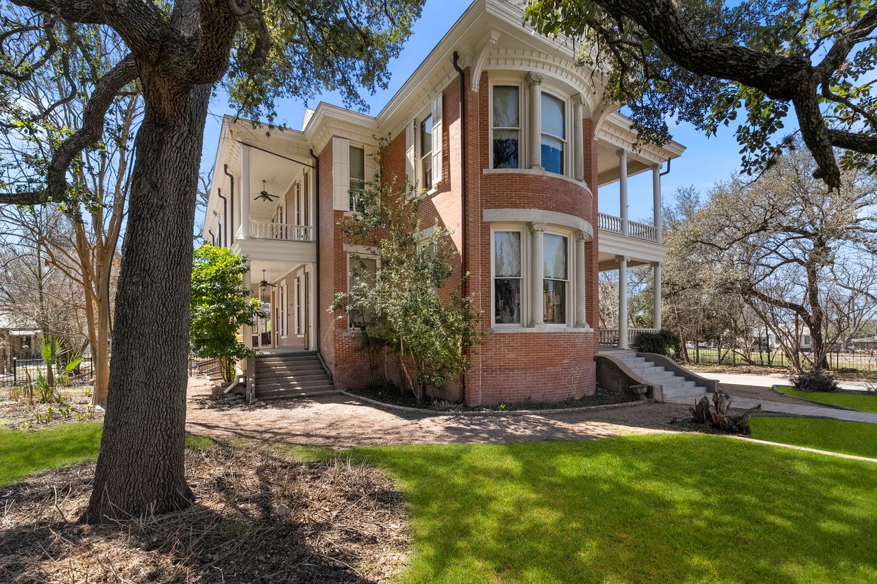 Corner view of the two-story red brick Dittlinger house with a rounded two-story bay window, two-tier white-columned porches and a large tree trunk in the foreground