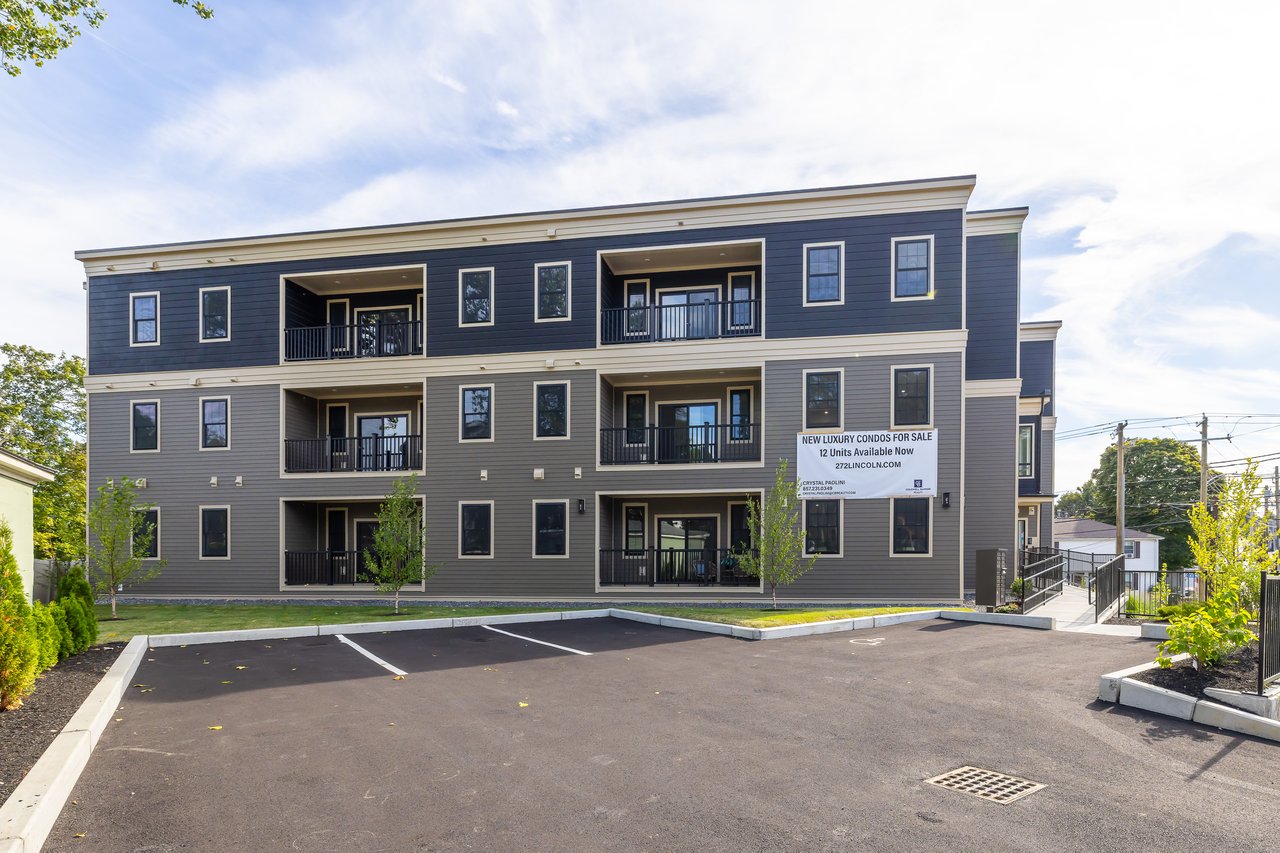 Three-story condominium building with recessed balconies, dark siding, and a parking area.