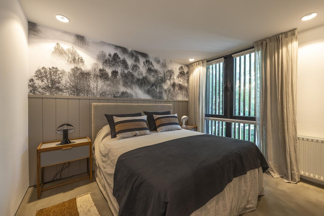 Furnished bedroom with a black-and-white forest mural behind the bed and tall curtained windows.