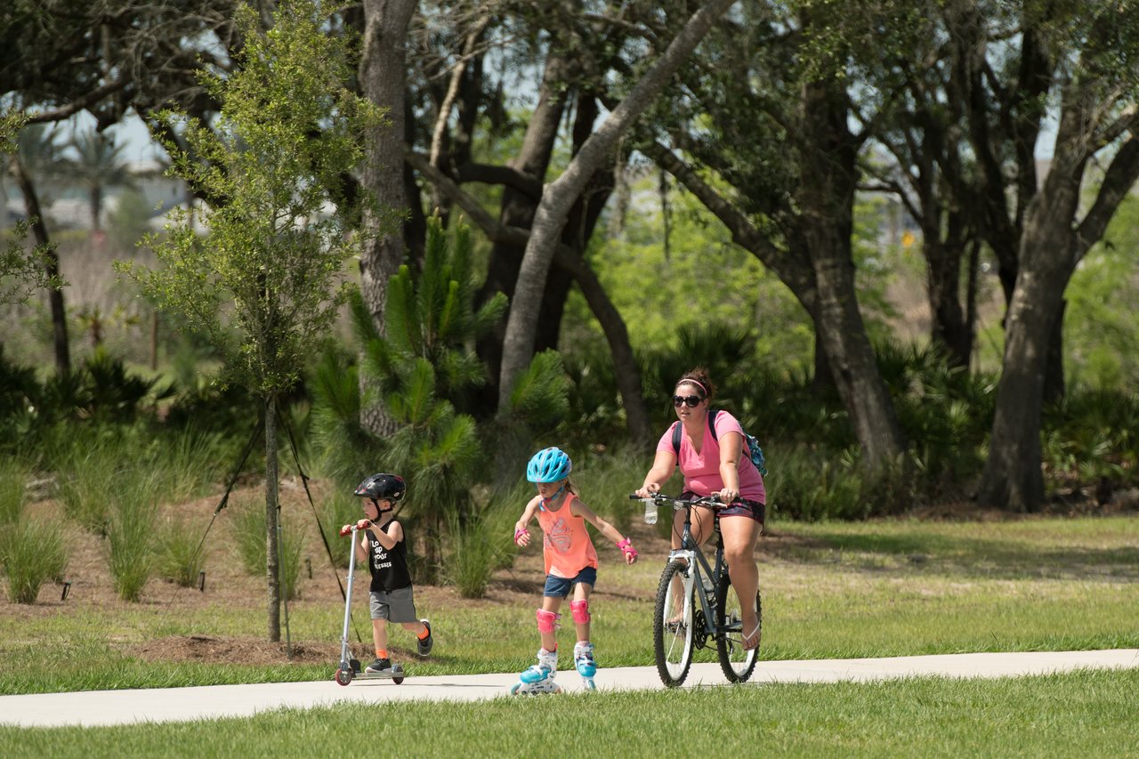 A woman bikes beside two children riding scooters on a path beneath leafy trees.