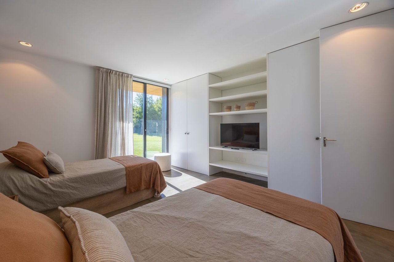 Modern bedroom with two neutral-toned beds, built-in television shelving, and sliding doors opening onto a lawn.