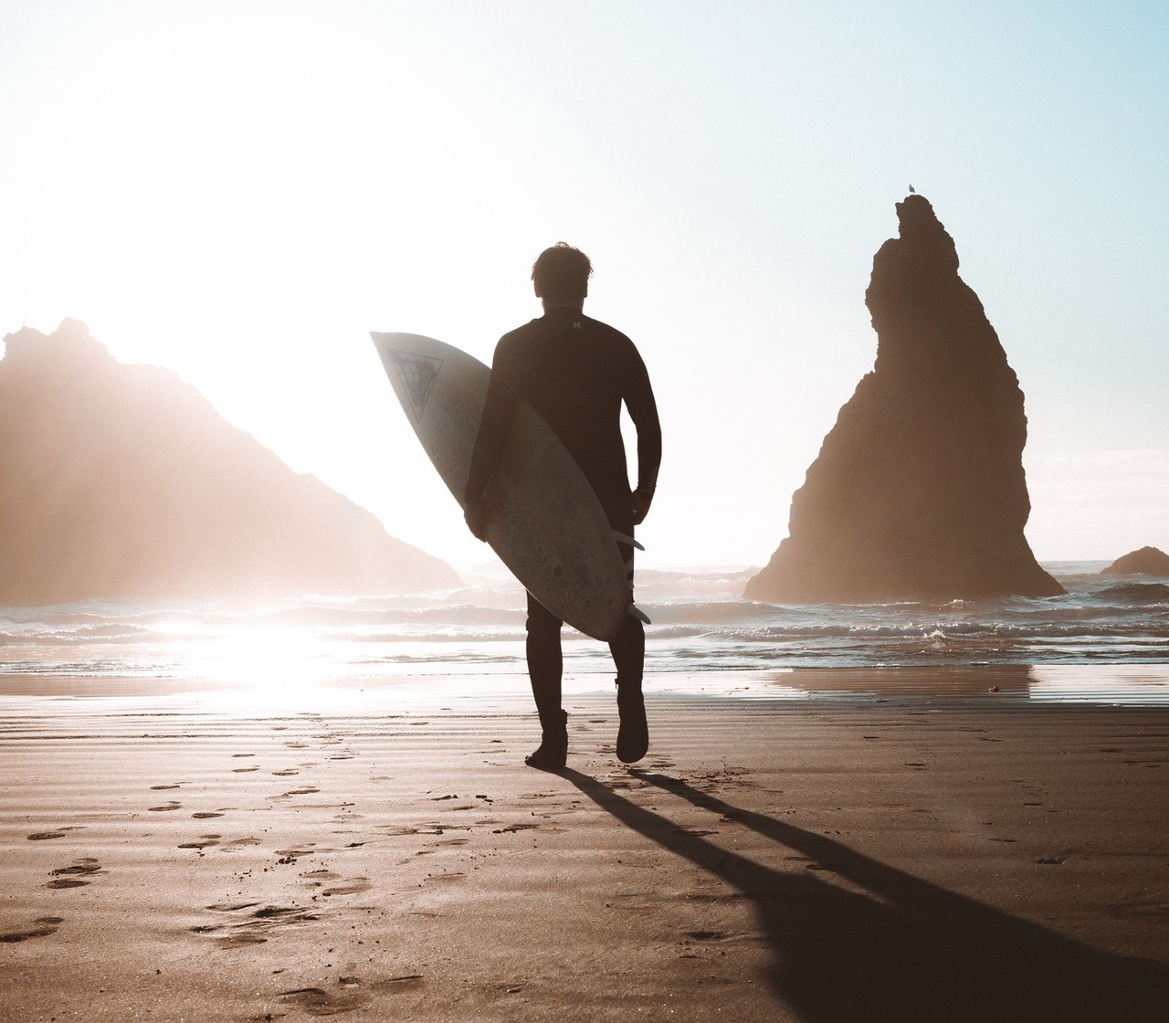 A surfer carrying a board walks toward the ocean between rocky sea stacks in warm backlight.