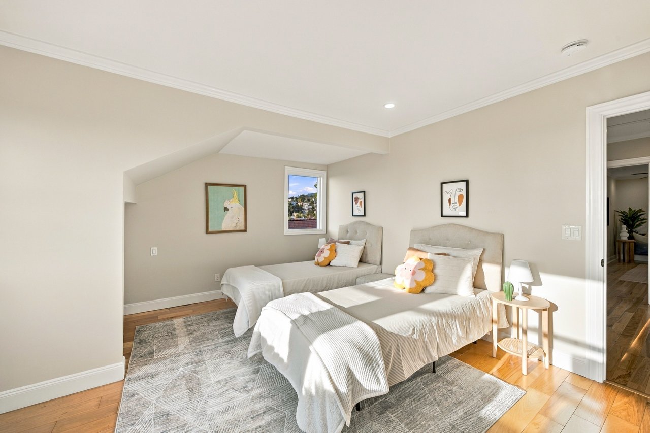 Staged bedroom with two upholstered beds, white bedding, and a window overlooking nearby homes.