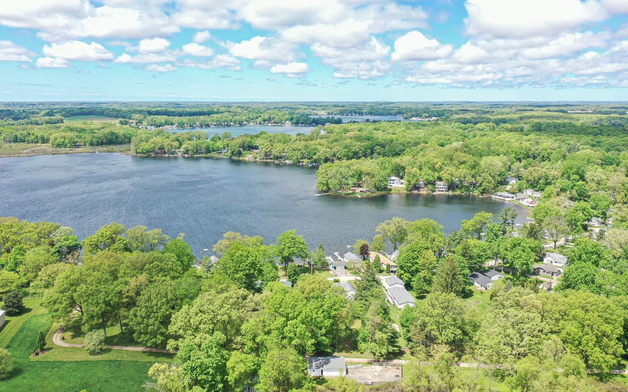 Aerial view of a lake and wooded shoreline homes beneath a partly cloudy sky.