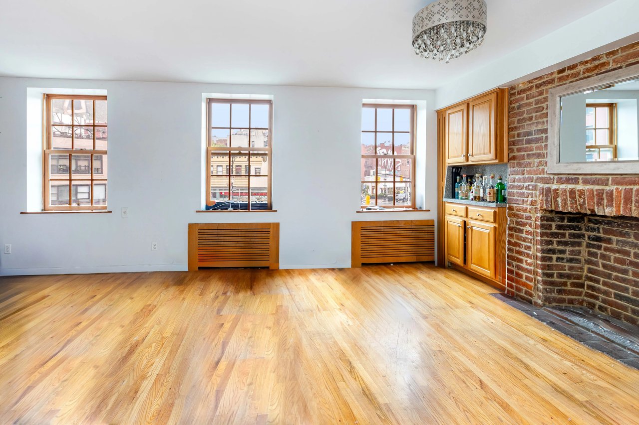 Vacant living room with hardwood floors, three tall windows, and an exposed-brick fireplace.