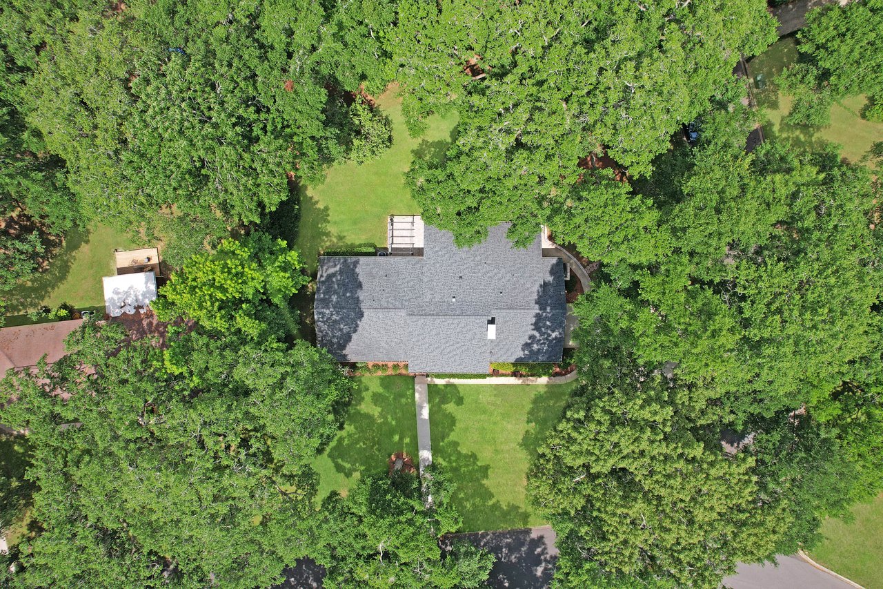 Aerial view of a house with a gray roof surrounded by lush green trees in a suburban area. A neat lawn and paved pathway are visible, creating a serene atmosphere.
