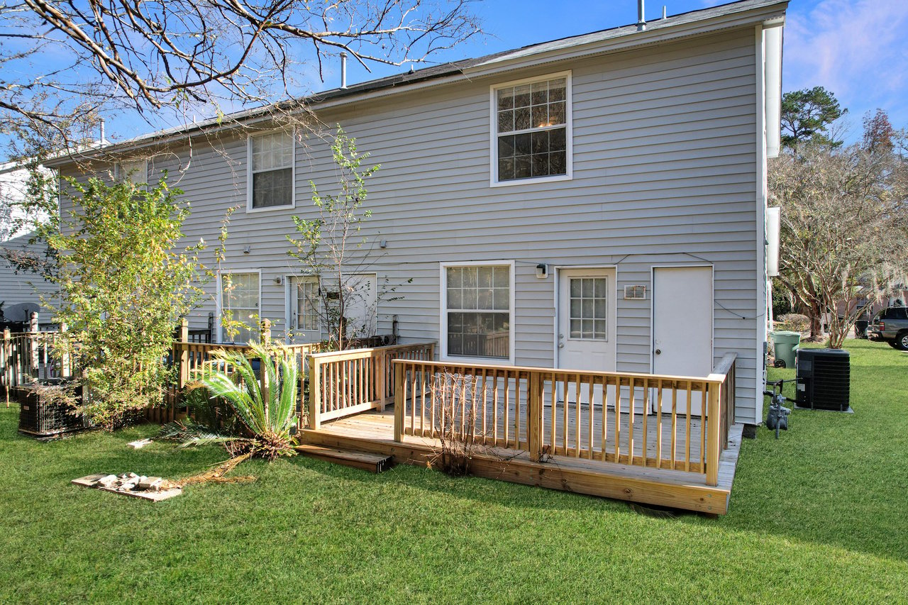 Two-story gray house with white trim, featuring a wooden deck and grassy backyard. Trees and plants surround the area, creating a peaceful ambiance.