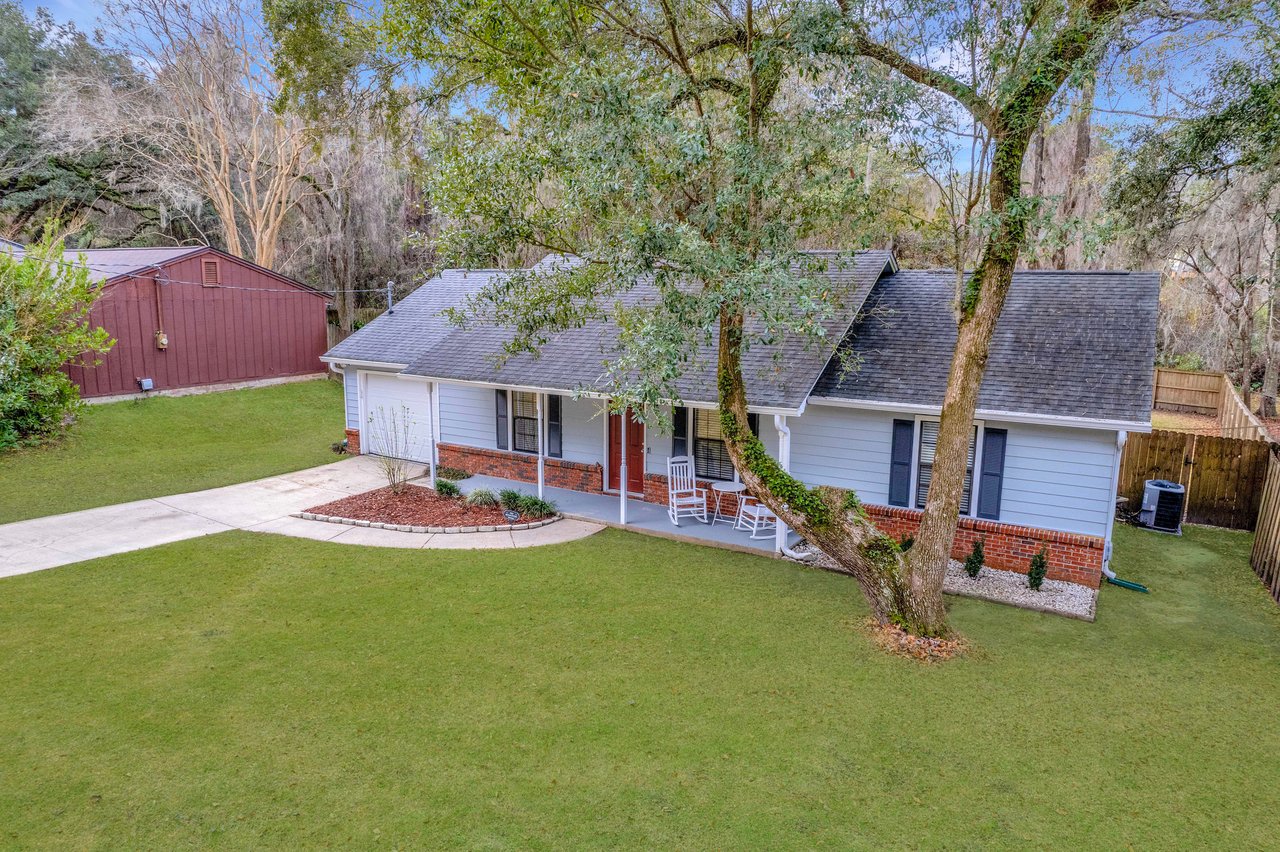 A quaint suburban house with a white and red brick exterior, wide porch, rocking chairs, and a large tree in the front yard. The scene is peaceful and inviting.