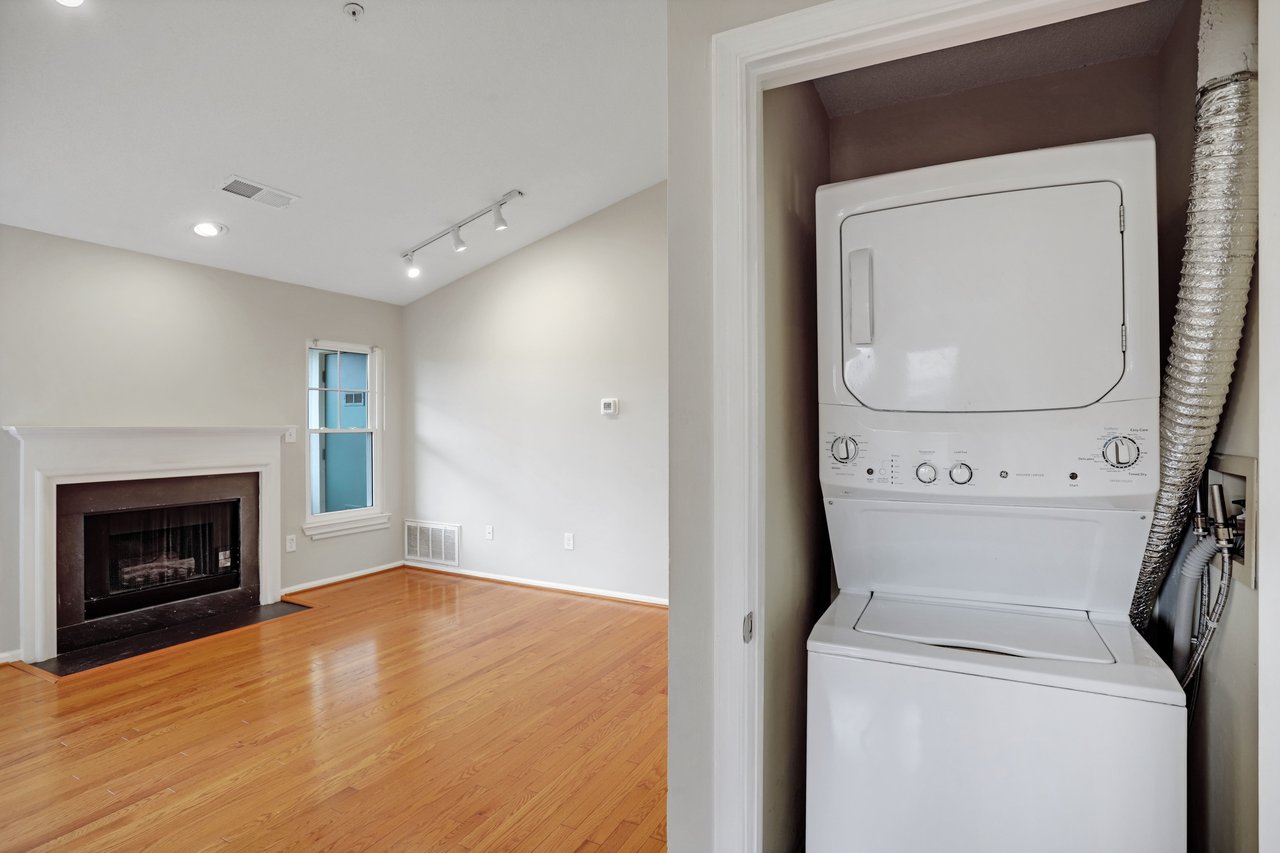 Vacant living room with hardwood floors and fireplace beside a closet with stacked washer and dryer.