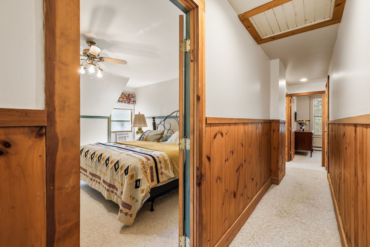 Furnished bedroom with a patterned bedspread beside a carpeted hallway with wood-paneled walls.