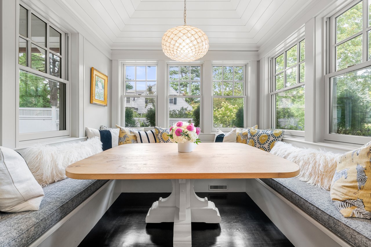 Dining nook with a light-wood table, built-in window seats, and tall windows overlooking trees.