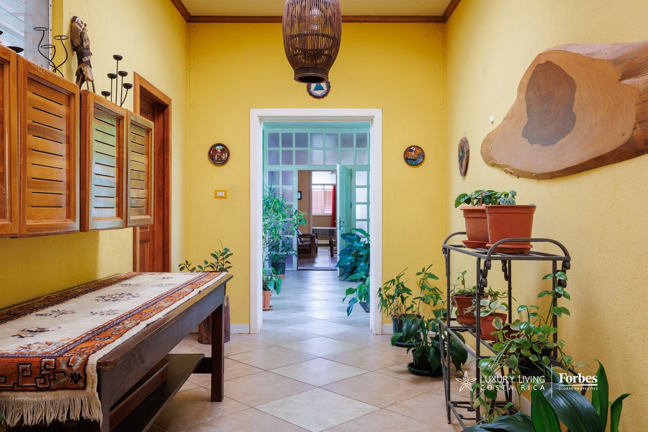Entryway with yellow walls, wooden shutters, a patterned console table, and plants beside an open doorway.